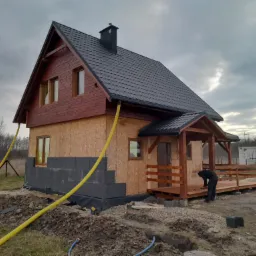 Surowy stan domu w trakcie budowy: drewniana konstrukcja szkieletowa z czarną dachówką, częściowo ocieplona fundamentami z szarych bloczków, z drewnianym tarasem i pracującą osobą na nim, widoczne...