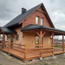 Dobra Stolarnia - Wznoszony dom szkieletowy z drewnianym tarasem i zadaszeniem, widoczna konstrukcja ścian z płyt OSB i częściowo wykończona drewniana elewacja, pracownik w trakcie montażu balustrady tarasu.