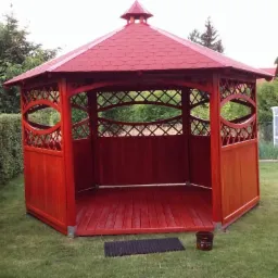 Czerwona, drewniana altana ogrodowa z dekoracyjnymi wycięciami i czerwonym dachem z gontu bitumicznego, ustawiona na trawniku obok żywopłotu i szklarni.