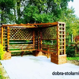 Drewniana pergola z ławkami i kratownicą, otoczona zielenią, z widocznym drzewkiem iglastym w donicy i brukowaną nawierzchnią