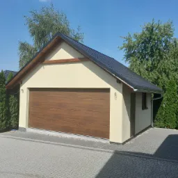 Nowoczesny, parterowy garaż z brązową bramą segmentową, elewacją w odcieniach beżu, ciemną dachówką i drewnianym wykończeniem pod dachem, otoczony kostką brukową i zielenią.