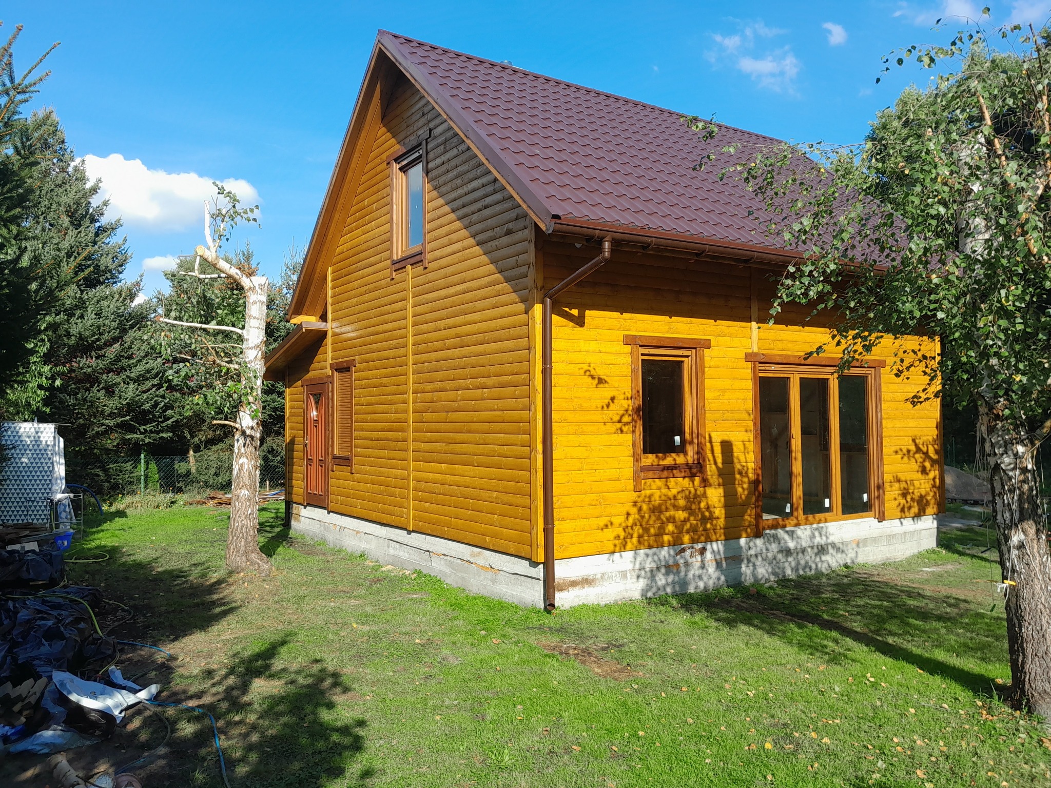 Żółty dom z drewnianą elewacją i brązowym dachem z blachodachówki, widoczny z perspektywy trawnika w słoneczny dzień.