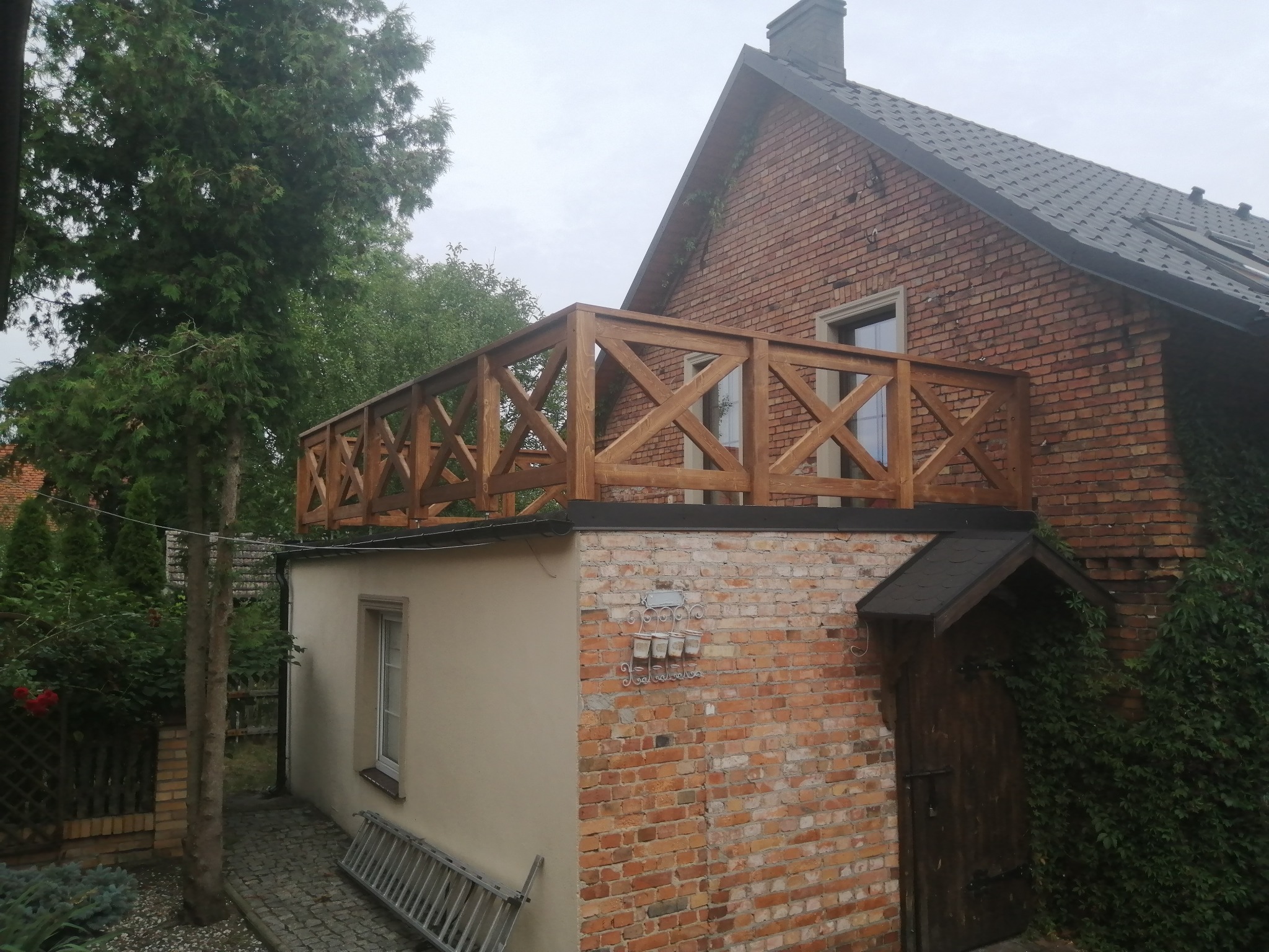 Zadaszony taras z drewnianą balustradą z krzyżowymi wzorami, umieszczony na płaskim dachu przybudówki z cegły, z drabina opartą o ścianę i zielenią w tle.