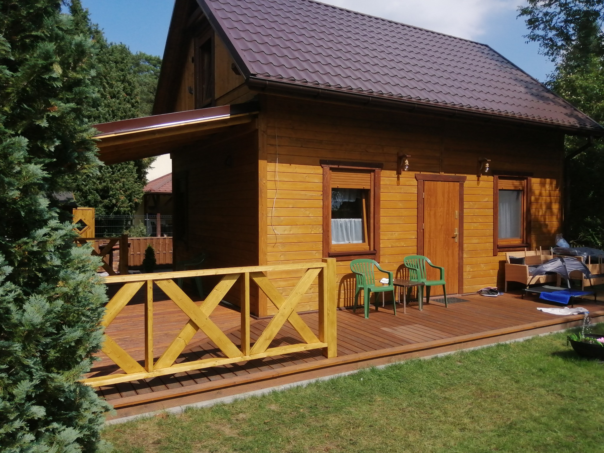 Drewniany domek z brązowym dachem i tarasem z drewnianą balustradą w kształcie krzyży, zielone krzesła plastikowe na tarasie, otoczenie trawnik i drzewa.