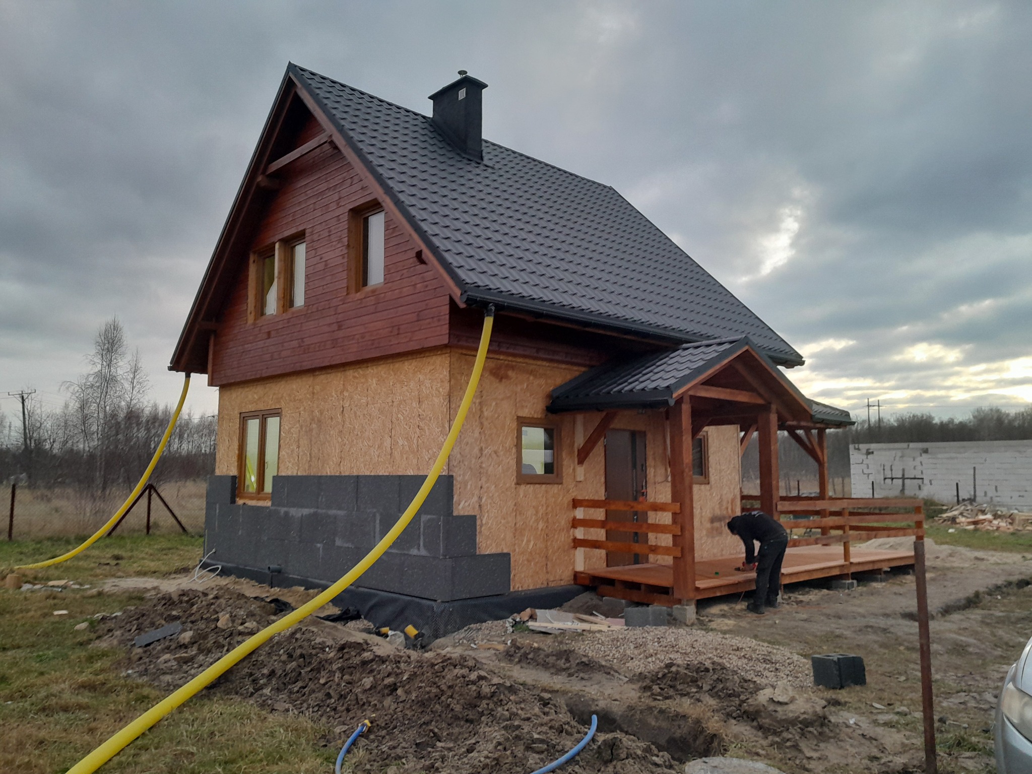 Surowy stan domu w trakcie budowy: drewniana konstrukcja szkieletowa z czarną dachówką, częściowo ocieplona fundamentami z szarych bloczków, z drewnianym tarasem i pracującą osobą na nim, widoczne...