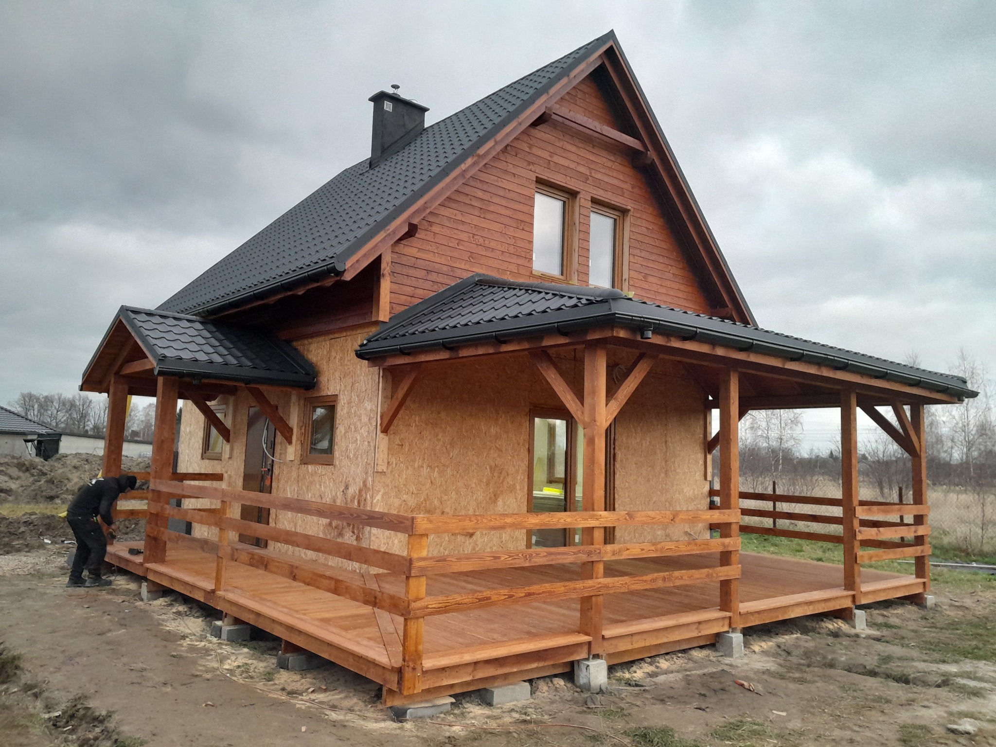 Wznoszony dom szkieletowy z drewnianym tarasem i zadaszeniem, widoczna konstrukcja ścian z płyt OSB i częściowo wykończona drewniana elewacja, pracownik w trakcie montażu balustrady tarasu.