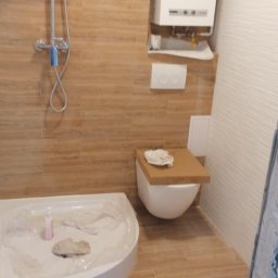 Pat-Bud - Wykończona łazienka z kabiną prysznicową, podwieszaną toaletą i piecem gazowym. Beżowe płytki imitujące drewno na ścianach i podłodze. Widoczny fragment opakowania paneli.