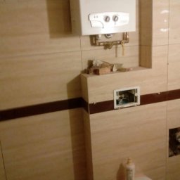 Pat-Bud - Biały piec gazowy na beżowych płytkach w łazience w trakcie prac. Widoczne narzędzia i niedokończona instalacja podtynkowa WC. Ujęcie z dołu.