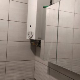 Pat-Bud - Biały piec gazowy z podłączoną rurą, zamontowany na ścianie z jasnych, kwadratowych płytek w łazience. Widoczne fragmenty lustra i niedokończone wykończenie ściany.