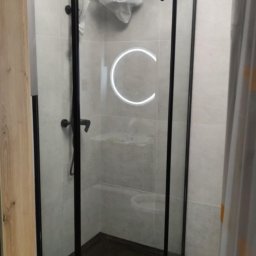 Pat-Bud - Nowoczesna kabina prysznicowa z czarnymi profilami i oświetleniem LED w kształcie okręgu, na tle jasnych płytek i drewnopodobnej podłogi. Widok zza framugi drzwi.