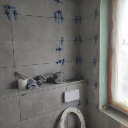 Pat-Bud - Szare płytki ścienne w trakcie układania w łazience, z użyciem niebieskich krzyżyków dystansowych i poziomicy laserowej. Widoczny fragment toalety i narzędzia.