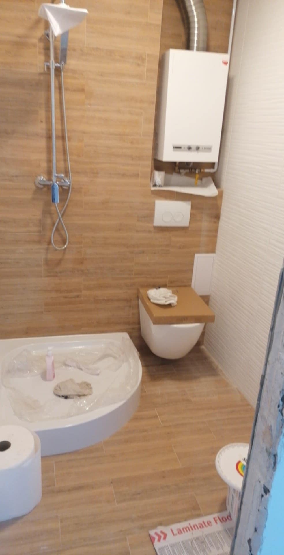 Wykończona łazienka z kabiną prysznicową, podwieszaną toaletą i piecem gazowym. Beżowe płytki imitujące drewno na ścianach i podłodze. Widoczny fragment opakowania paneli.