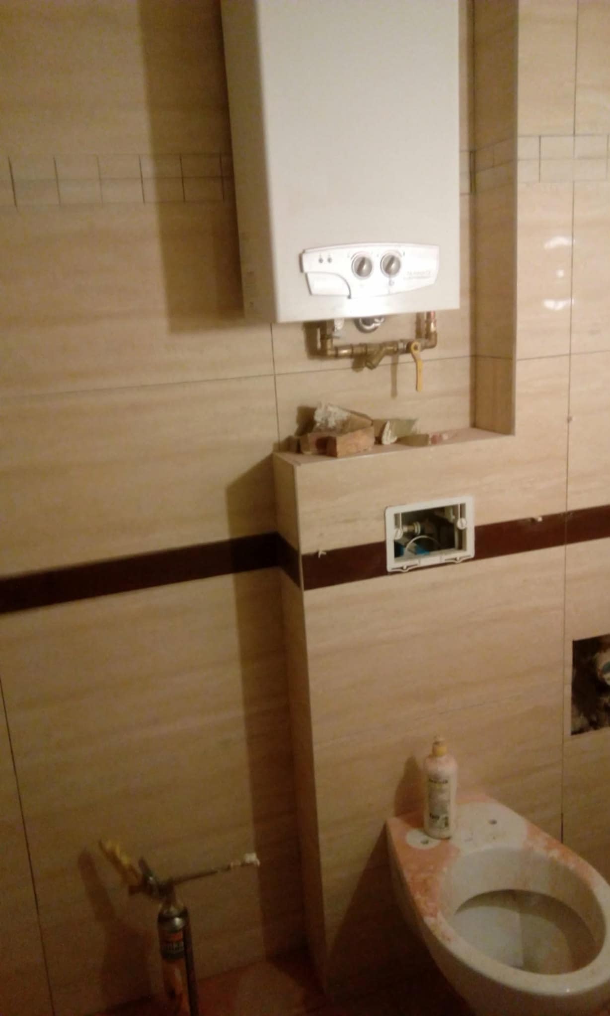 Biały piec gazowy na beżowych płytkach w łazience w trakcie prac. Widoczne narzędzia i niedokończona instalacja podtynkowa WC. Ujęcie z dołu.