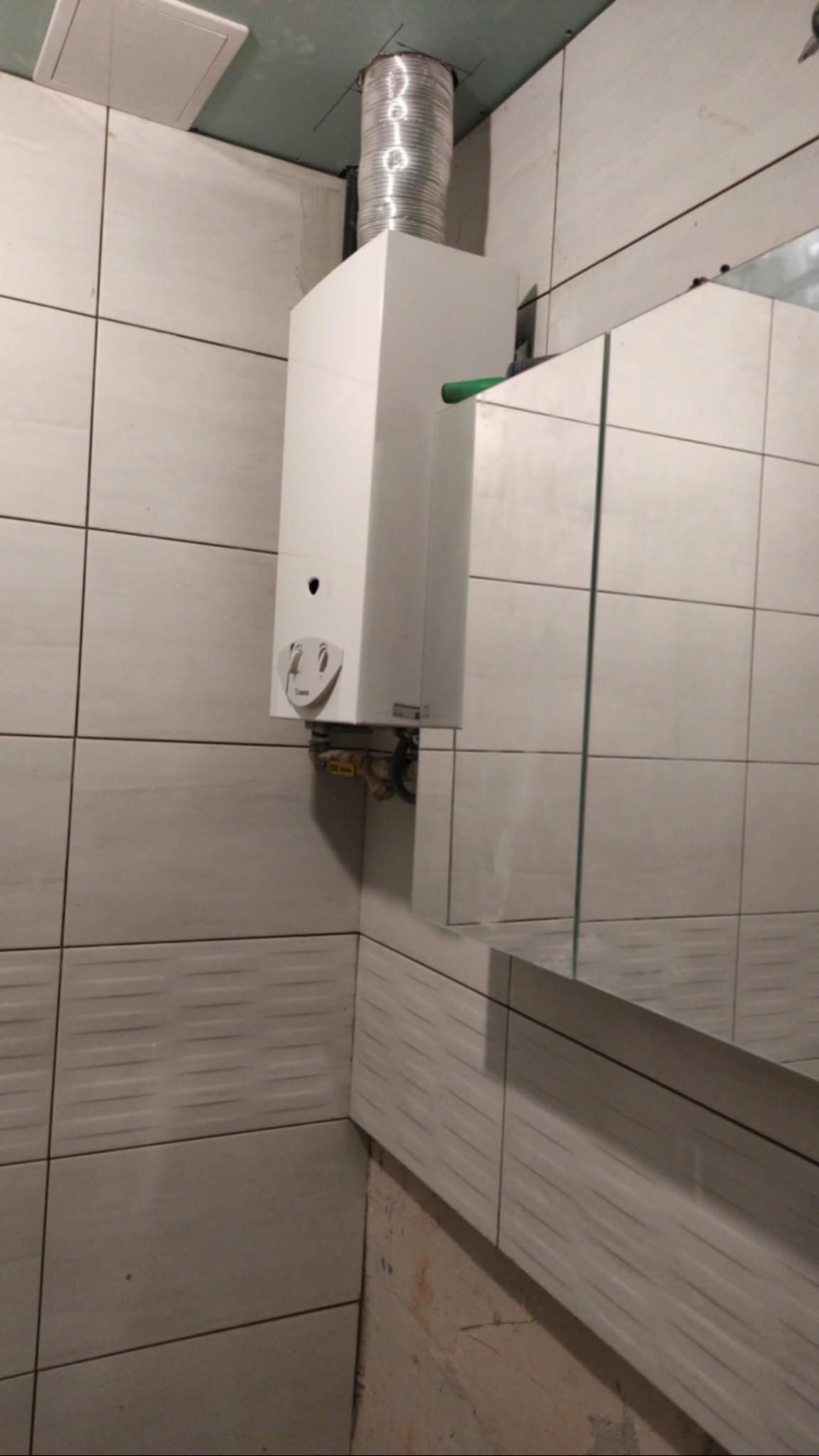Biały piec gazowy z podłączoną rurą, zamontowany na ścianie z jasnych, kwadratowych płytek w łazience. Widoczne fragmenty lustra i niedokończone wykończenie ściany.