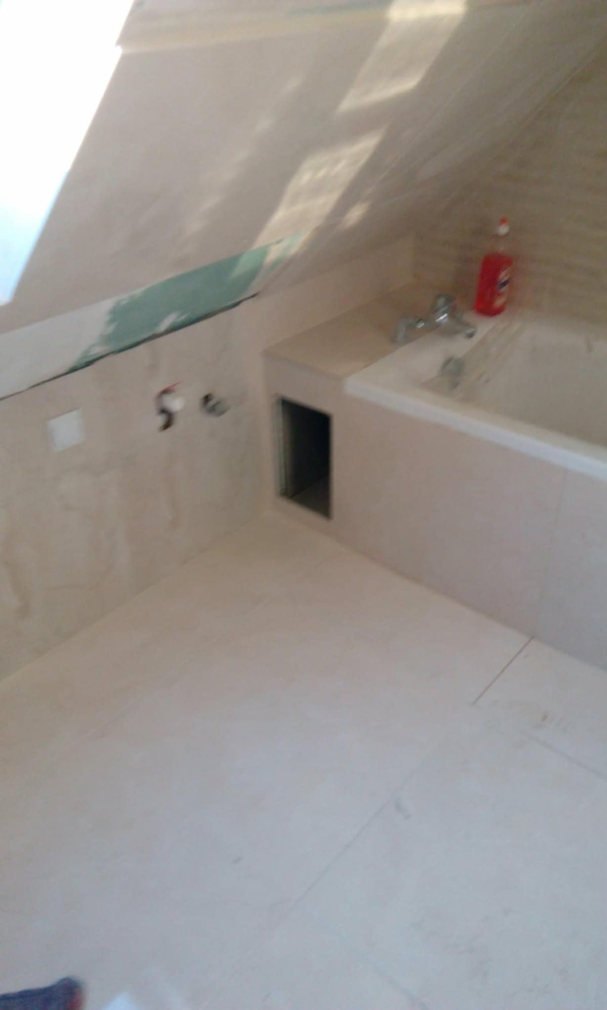 Wykończona łazienka na poddaszu z beżowymi płytkami, wanną i małym otworem rewizyjnym. Widoczne fragmenty niedokończonych prac na ścianie.