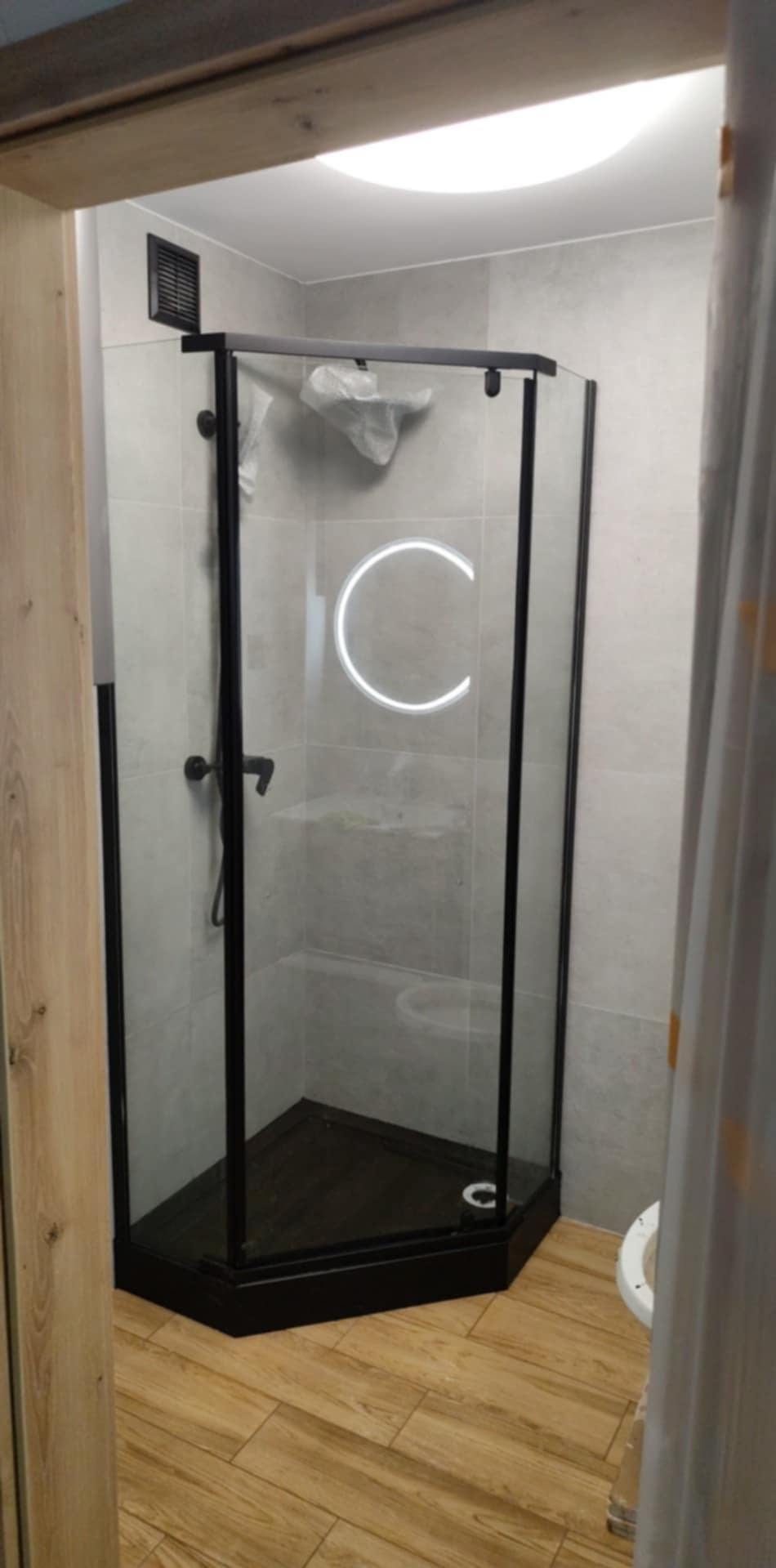 Nowoczesna kabina prysznicowa z czarnymi profilami i oświetleniem LED w kształcie okręgu, na tle jasnych płytek i drewnopodobnej podłogi. Widok zza framugi drzwi.