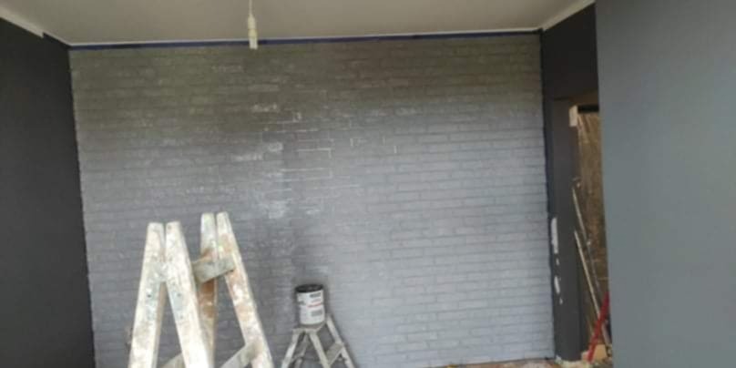 Ściana z efektem cegły pomalowana na szaro, drabina malarska i puszka farby. Widoczne ślady po taśmie malarskiej na suficie.