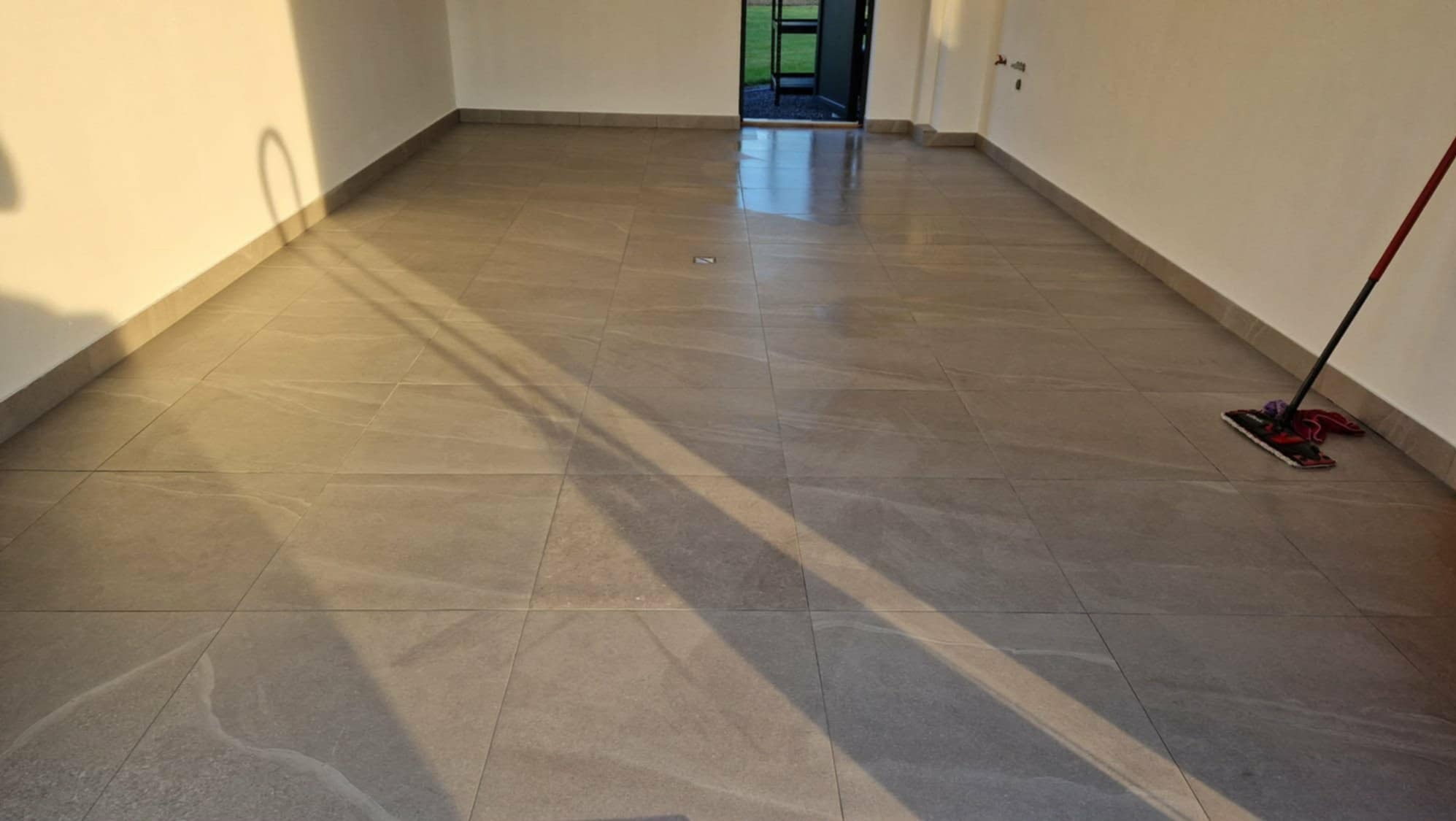 Ułożona, szara podłoga z płytek w pustym pomieszczeniu, widoczny mop przy ścianie. Naturalne światło podkreśla strukturę płytek i czystość wnętrza.