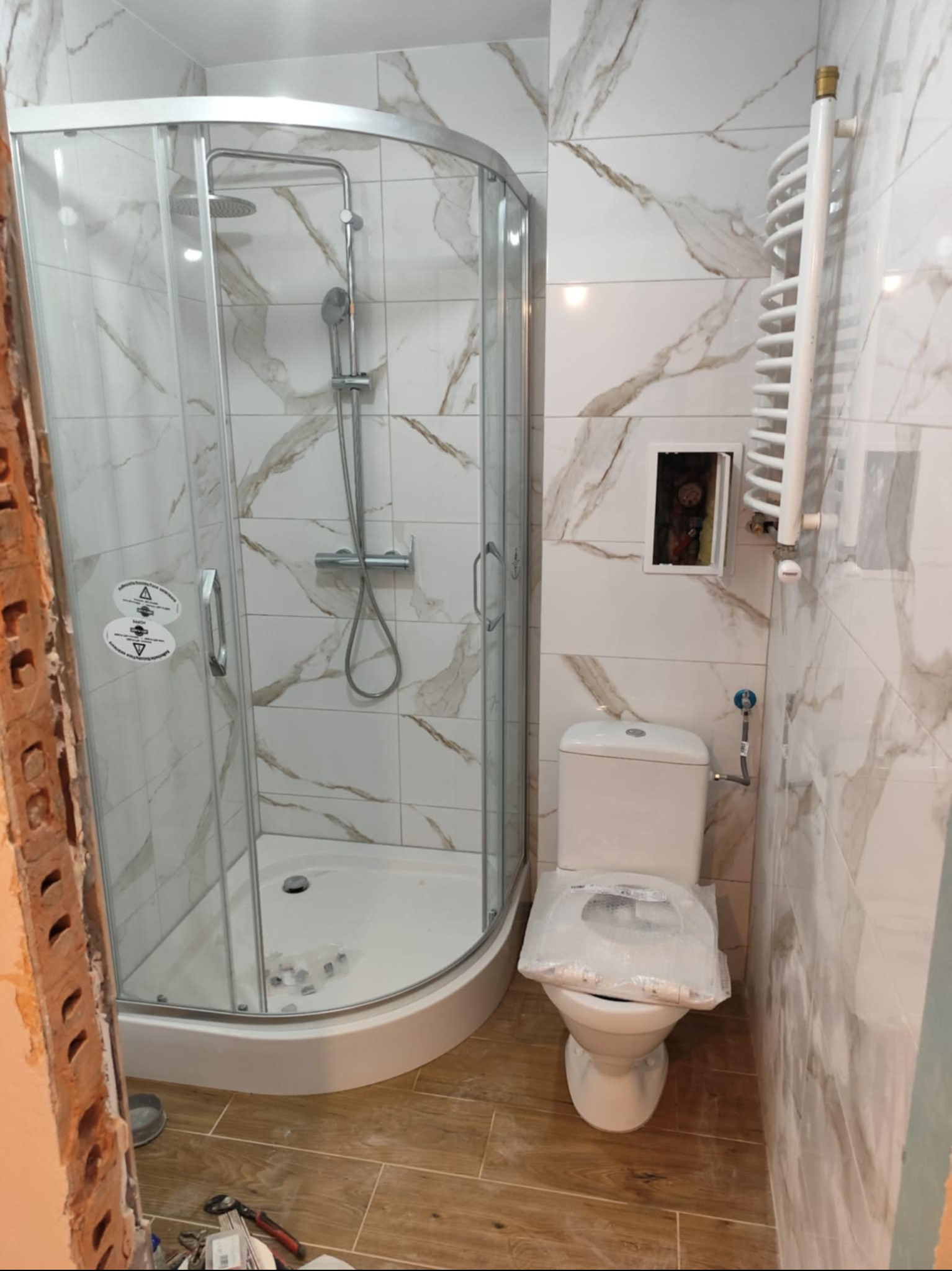 Kabina prysznicowa z brodzikiem, toaleta i grzejnik na tle marmurowych płytek w łazience po remoncie w Gliwicach. Widoczne narzędzia.