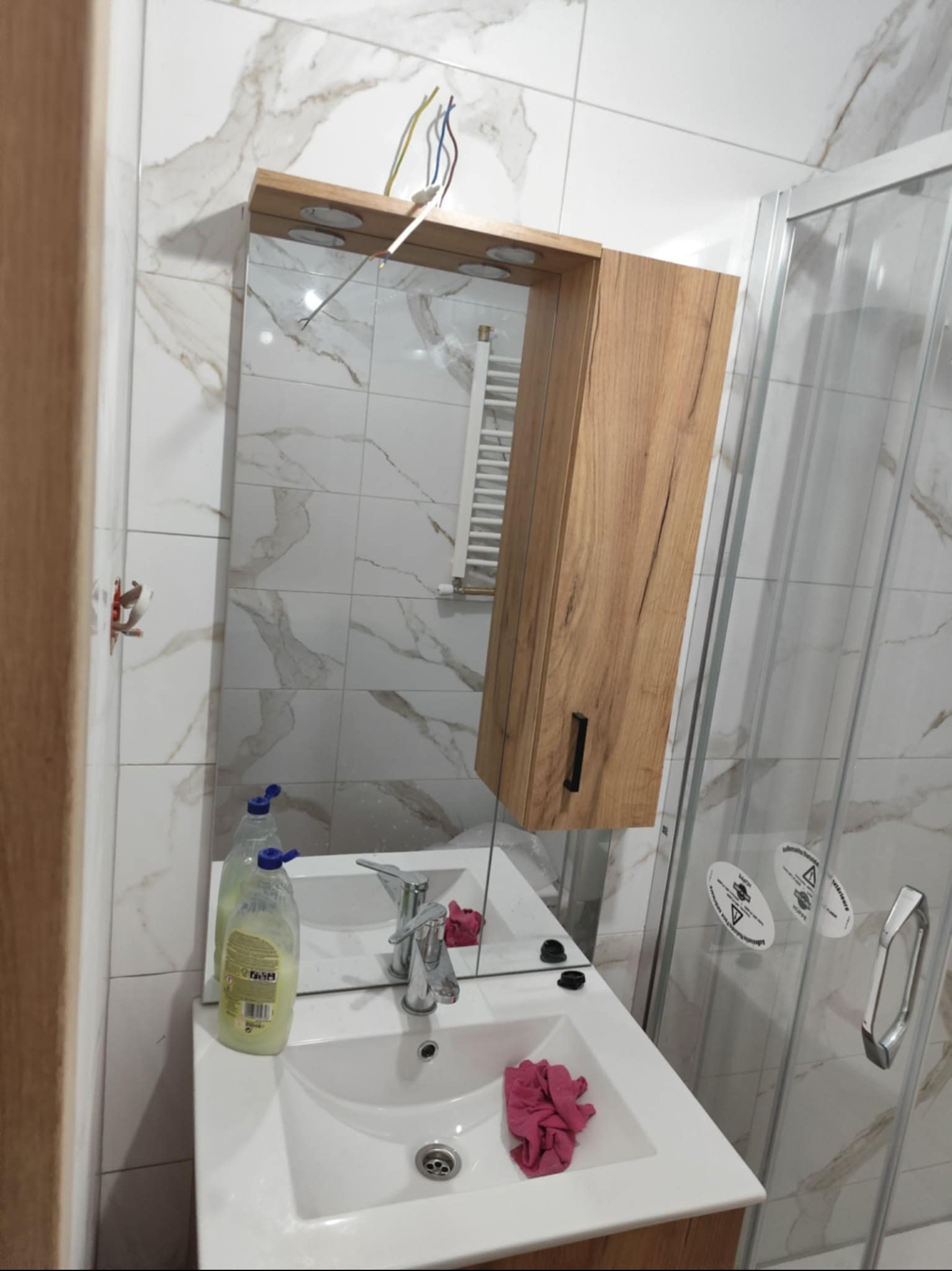 Łazienka po remoncie: białe płytki z marmurowym wzorem, umywalka z szafką i lustrem z oświetleniem. Widoczne wystające przewody elektryczne i kabina prysznicowa.