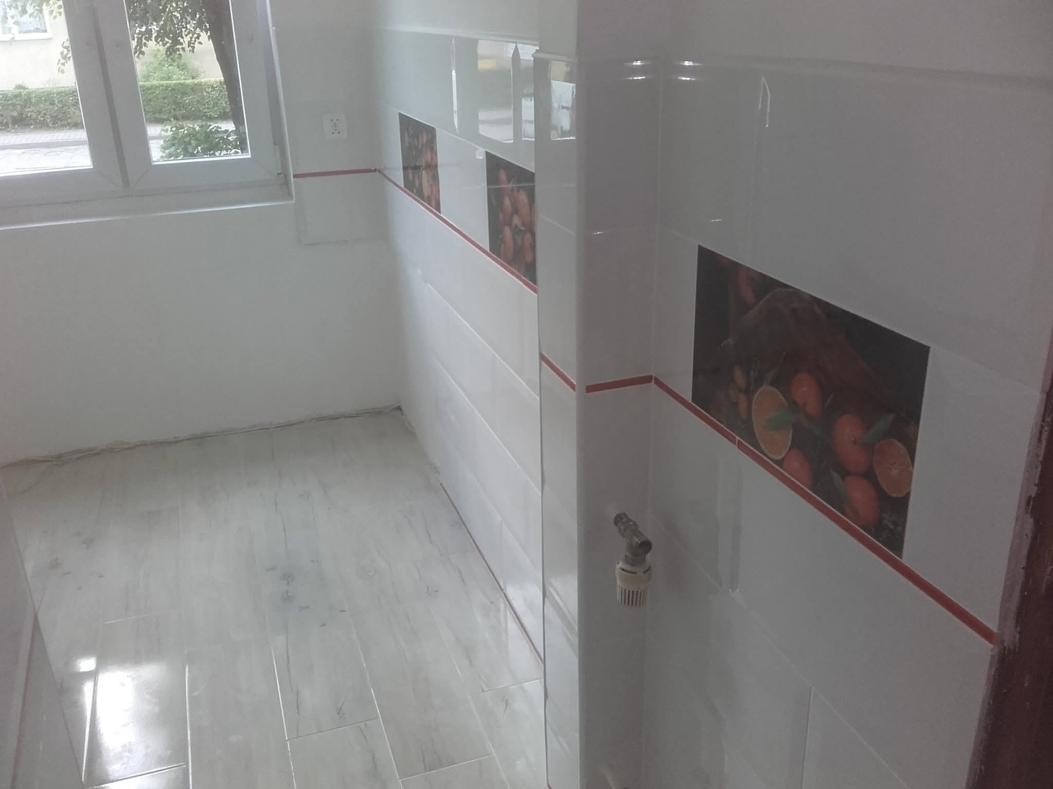 Wykończona łazienka z białymi płytkami, dekoracyjnymi panelami z motywem owoców i jasną podłogą. Widoczny fragment okna i instalacja wodna.