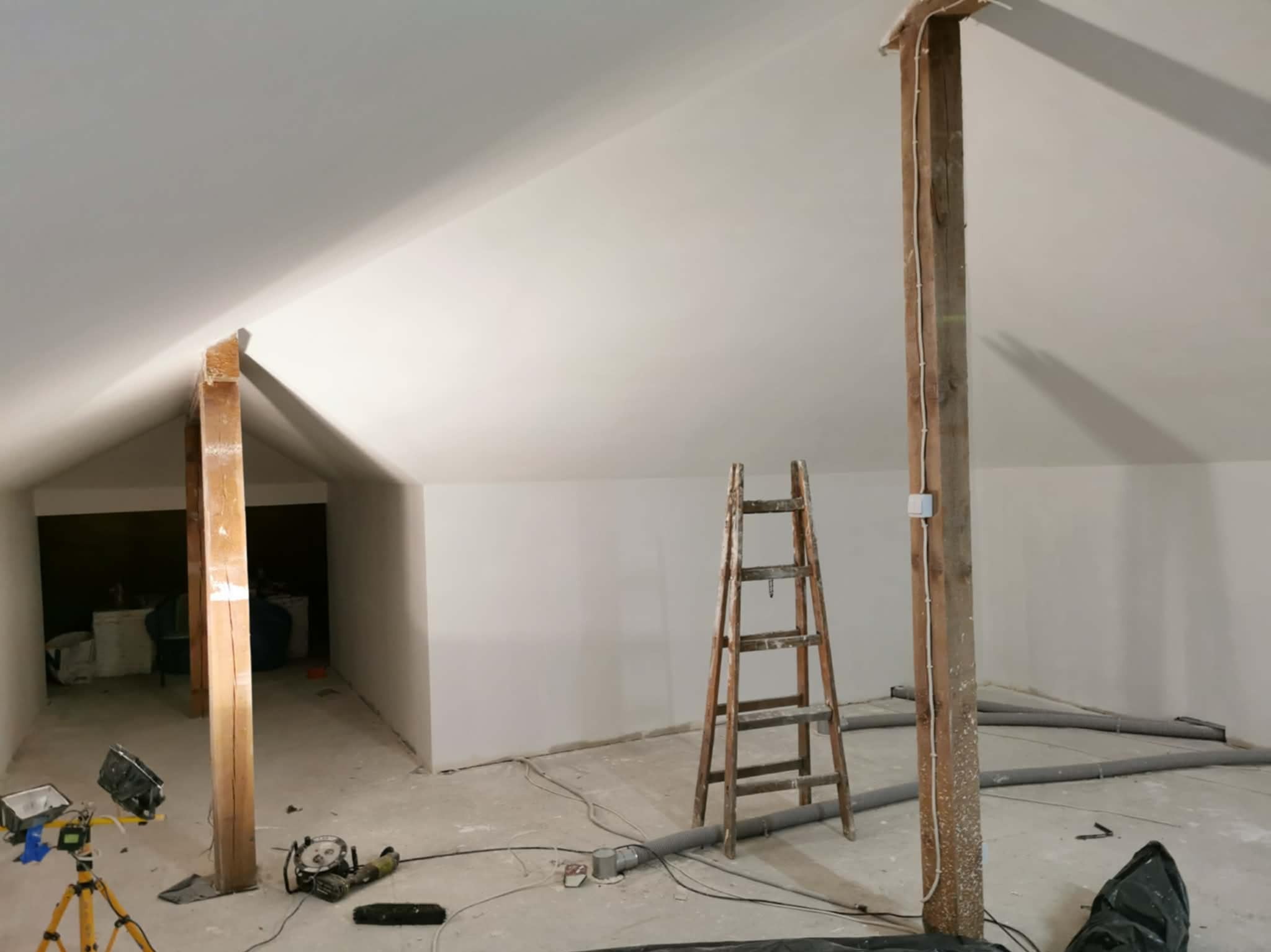 Poddasze w trakcie prac wykończeniowych: drewniane słupy, drabina, przewody wentylacyjne na podłodze, lampa budowlana. Ściany i sufit pomalowane na biało.
