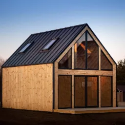 Nowoczesny, parterowy budynek z drewnianą elewacją i dużym przeszkleniem szczytowym, kryty blachą na rąbek stojący, z dwoma oknami dachowymi, na tle zachodzącego słońca.
