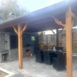 Zadaszona konstrukcja ogrodowa z grillem murowanym z szarej cegły, drewnianymi słupami o dekoracyjnych wspornikach, zasłonięta ażurową ścianką, z kompletem mebli ogrodowych i szarą kostką brukową...