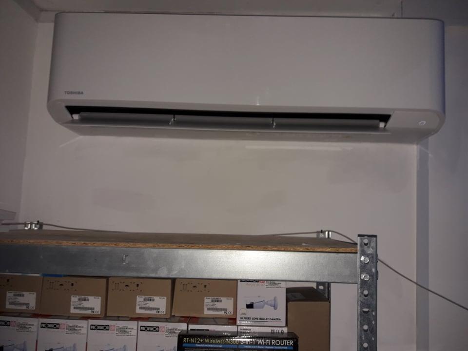 Biała jednostka wewnętrzna klimatyzatora Toshiba zamontowana na ścianie, widoczna półka metalowa z kartonami poniżej.