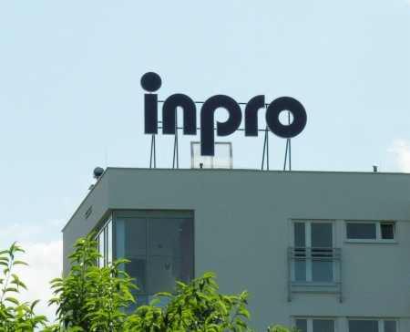 Budynek biurowy z dużym, czarnym napisem 'inpro' na dachu, widoczny z dołu przez korony drzew.