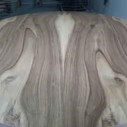 Okrągły blat stołu z drewna o wyraźnym, symetrycznym usłojeniu, ukazujący naturalne wzory i odcienie brązu, z widocznymi elementami warsztatu w tle.