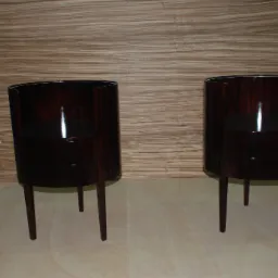 Dwa identyczne, okrągłe stoliki nocne z ciemnego drewna z dwiema szufladami, stojące na drewnianej podłodze na tle drewnianej ściany.