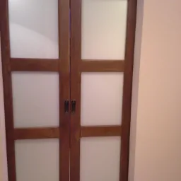 Dwuskrzydłowe przesuwne drzwi z drewna o ciepłym odcieniu, z matowym szkłem w prostokątnych panelach, zamontowane w jasnym pomieszczeniu.