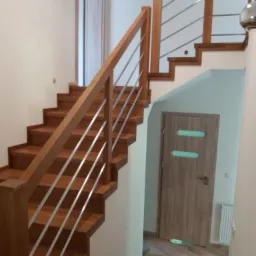 Nowoczesne schody z drewna z metalowymi barierkami, widok z dołu na tle białych ścian i drewnianych drzwi z podłużnymi przeszkleniami.