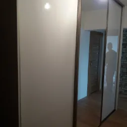 Biała szafa przesuwna z lustrzanymi drzwiami, widoczny fragment wnętrza z drewnianą podłogą i drzwiami oraz odbicie sylwetki człowieka.