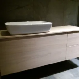 Nowoczesna łazienka z wiszącą szafką pod umywalkę z jasnego drewna i białą, prostokątną umywalką nablatową na tle ciemnoszarej ściany.