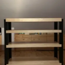 Prosta, drewniana konstrukcja regału z czterema półkami, wsparta na czarnych, metalowych nogach. Widoczny parkiet na podłodze i fragment komody w tle.