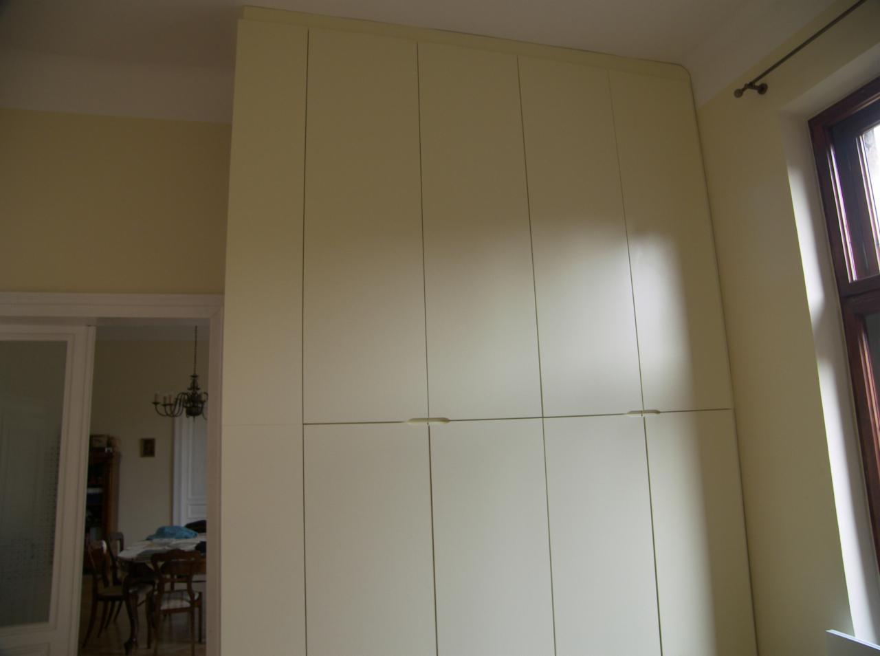 Wysoka, kremowa zabudowa meblowa od podłogi do sufitu, z widocznymi minimalistycznymi uchwytami, na tle jasnych ścian i fragmentu pokoju z żyrandolem w tle.