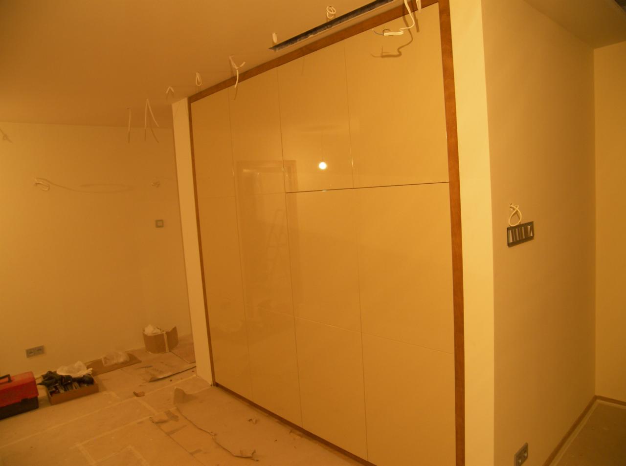 Beżowa, lakierowana zabudowa szafy wnękowej z drewnianymi obramowaniami, widoczna w trakcie montażu, z wystającymi kablami, narzędziami i materiałami budowlanymi w pomieszczeniu.