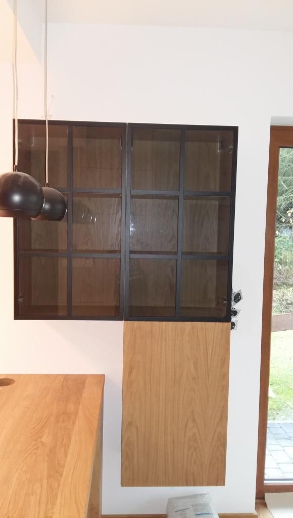 Część zabudowy kuchennej: wiszące szafki przeszklone z czarnymi ramami nad drewnianym blatem i szafką, widoczne elementy instalacji elektrycznej przy oknie.