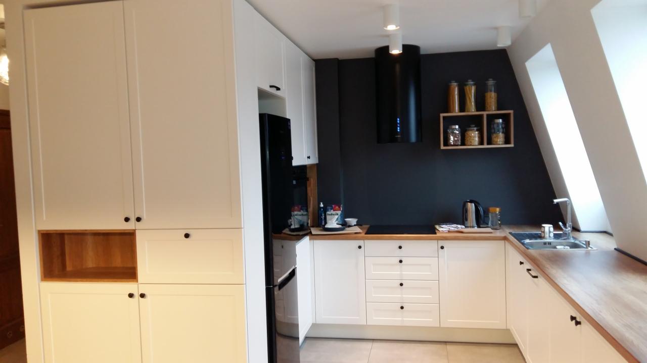 Biała kuchnia z drewnianym blatem w mieszkaniu na poddaszu, czarny okap nad płytą grzewczą, półka z słoikami z makaronem na ciemnej ścianie.