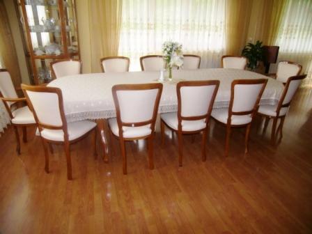 Jadalnia z owalnym stołem nakrytym białym obrusem z koronkowym wykończeniem i kompletem krzeseł z drewnianymi ramami i jasnym obiciem, w tle przeszklona witryna z porcelaną.