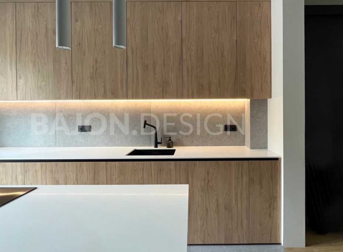 Nowoczesna kuchnia z drewnianymi szafkami, podświetleniem LED pod górnymi szafkami i czarną baterią umywalkową, widoczne logo 'BAJON.DESIGN' na ścianie.