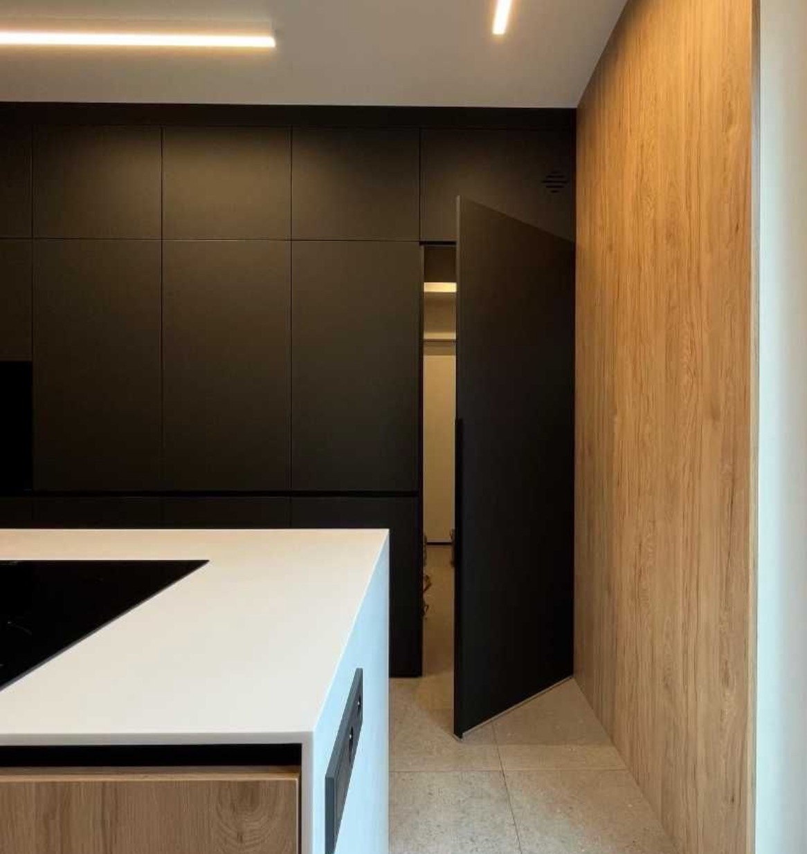 Nowoczesna kuchnia z czarnymi szafkami, białą wyspą i ukrytymi drzwiami, wykończona drewnianą okładziną ścienną.