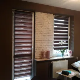 Dwa okna z zamontowanymi roletami dzień-noc w brązowo-białej kolorystyce, pomiędzy oknami fragment ściany z cegły dekoracyjnej, na parapecie storczyk w doniczce, pluszowa zabawka i szklane wazony.