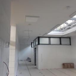 Remonter - Wnętrze w trakcie prac wykończeniowych: białe ściany z glazurą, sufit z panelami LED i oknami dachowymi, widoczne kable i zabezpieczenia podłogi.