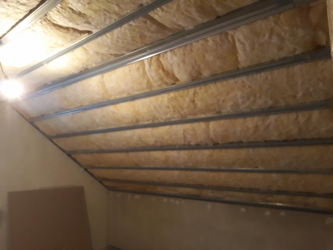 Izolacja poddasza z wełny mineralnej zamontowana między metalowymi profilami, widoczne fragmenty ścian i płyta kartonowo-gipsowa w rogu.