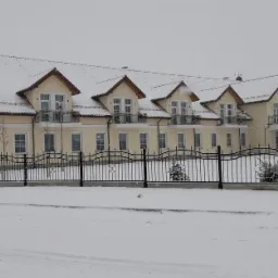 Nowo wybudowany budynek wielorodzinny z balkonami, pokryty śniegiem, ogrodzony czarnym, metalowym płotem w zimowej scenerii.