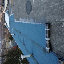 Nowe pokrycie dachu papą termozgrzewalną w kolorze niebieskim, widoczna rolka papy z nadrukiem liści klonu oraz fragment miasta w tle.