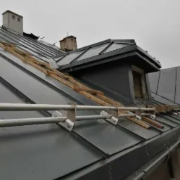 Szary dach z blachy trapezowej z zabezpieczeniem przeciwśniegowym i elementami drewnianymi przygotowanymi do montażu, widok z dołu na tle pochmurnego nieba.